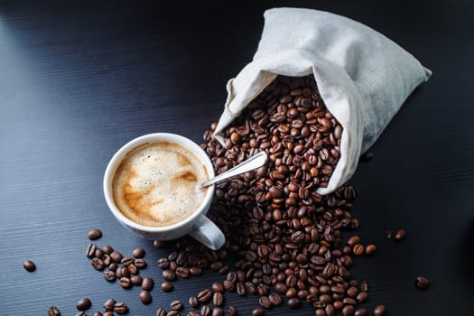 Cup of espresso and coffee beans scattered from a canvas bag on black wooden table background.