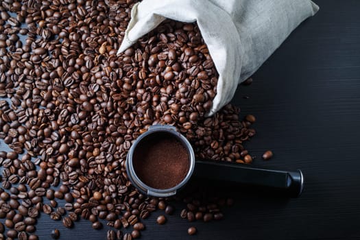 Coffee beans scattered from a canvas bag and holder from an espresso machine against a black wooden table background.