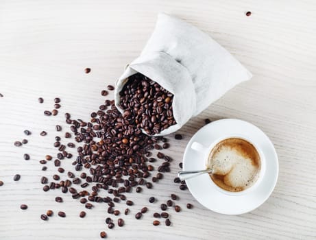 Espresso, coffee cup, saucer, spoon, and coffee beans scattered from a canvas bag on light oak wooden table. Top view.