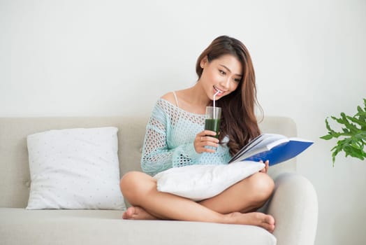 Pretty young asian woman drinking green fresh vegetable juice or smoothie from glass at home