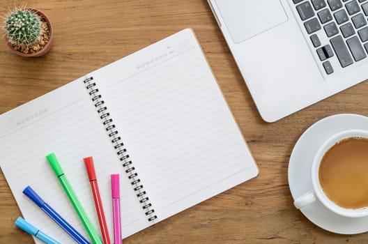 Wooden desk table with notebook, computer laptop, color pens, cactus and cup of coffee. Top view with copy space, flat lay.