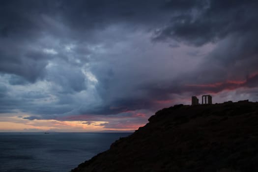 Cape Sounion, Attica, Greece - silhouette of the temple of Poseidon in the dusk, with distant ships and open sea background, under heavy grey, red and purple clouds.