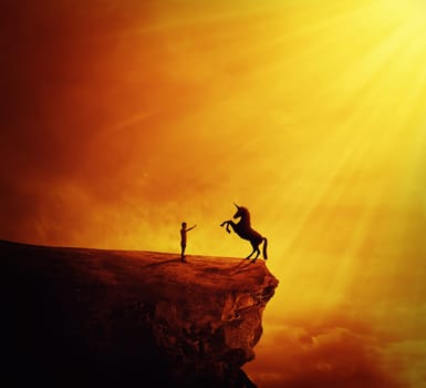 Idyllic view with a boy standing at the edge of a cliff chasm trying to tame a wild unicorn. Begining of a new friendship, fearless symbol