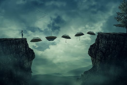 Young boy standing on the peak of a cliff try to pass over valley to another side on a path way of flying umbrellas. The pursuit of happiness and success concept.