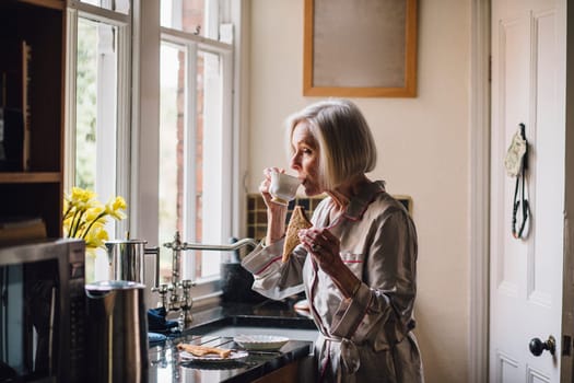Senior woman is standing alone in her kitchen in her dressing gown, eating toast and dirnking tea.