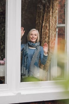 Senior woman is waving goodbye from a window in her home.