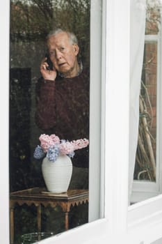 Senior man is looking out from his window into the street and is reporting a neighborhood crime on the phone.