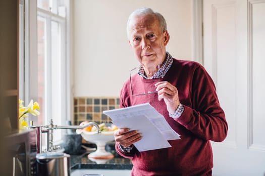 Senior man is standing in the kitchen of his home with bills in his hand, looking at the camera with a worried expression.