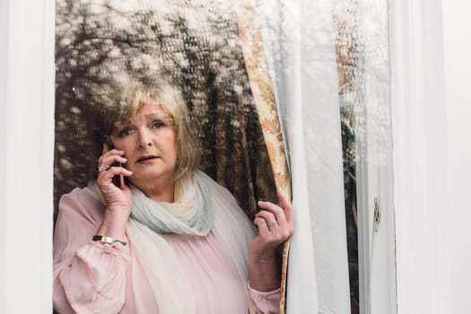 Senior woman is reporting a neighborhood crime to the police on the phone. She is looking out her window.