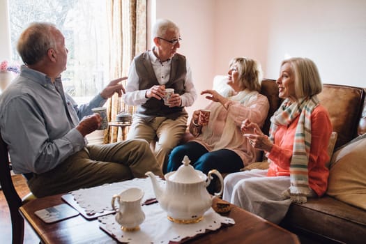 Group of seniors are sitting in the living room of a home together, enjoying a pot of tea and a chit-chat together.