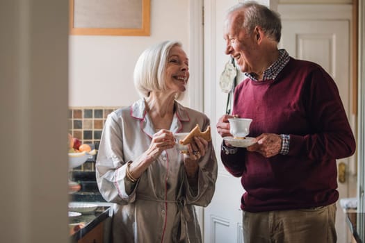 Senior couple are enjoying breakfast and talking in the kitchen. They are both drinking tea.