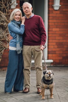 Happy senior couple are posing for the camera outside of their house, with their pet dog on a leash.
