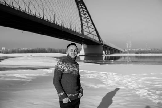 man in warm clothes with a beard is walking on the street in the winter in a warm sunny day at the river bridge in the background