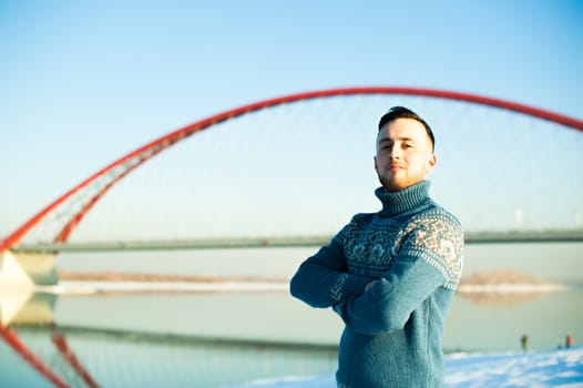 man in warm sweater with a beard is walking on the street in the winter in a warm sunny day by the river on a background of red bridge