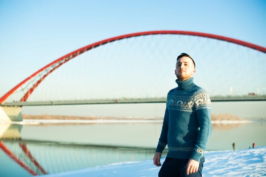 man in warm sweater with a beard is walking on the street in the winter in a warm sunny day by the river on a background of red bridge