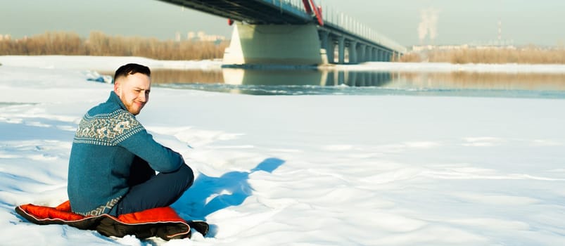 man in warm clothes with a beard is walking on the street in the winter in a warm sunny day at the river bridge in the background, sits in the snow