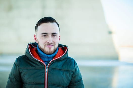 man in warm clothes with a beard is walking on the street in the winter in a warm sunny day at the river bridge in the background
