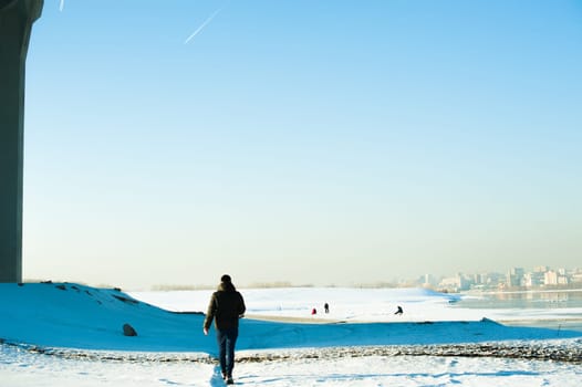 man in warm clothes with a beard is walking on the street in the winter in a warm sunny day at the river against the backdrop of the city