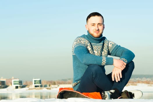 man in warm sweater with a beard is walking on the street in the winter in a warm sunny day at the river against the backdrop of the city, sitting in the snow on the beach with a view of the opposite shore