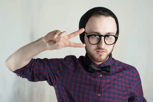studio portrait of a young man hipster. Guy with beard wearing glasses, hat and a plaid shirt, looking into the camera, showing two fingers