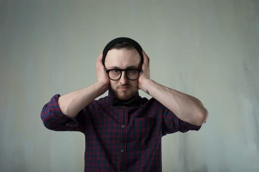 studio portrait of a young man hipster. Guy with beard wearing glasses, hat and a plaid shirt, grabbed his head