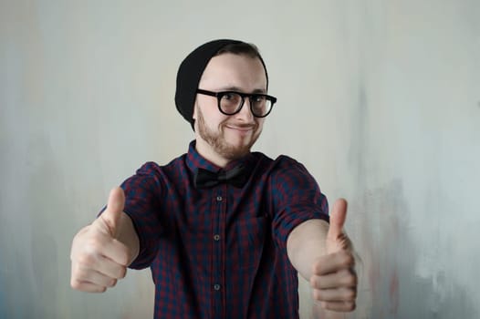 studio portrait of a young man hipster. Guy with beard wearing glasses, hat and a plaid shirt, showing a thumbs up