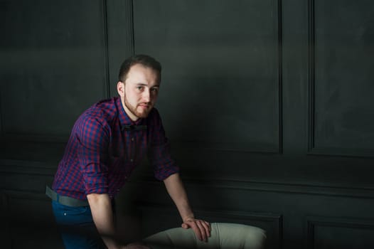 studio portrait of a young man on a dark wall background in a burgundy plaid shirt and blue jeans. hipster man, dressed stylishly, with a beard, in a room with an armchair