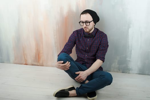 studio portrait of a young man hipster. Guy with beard wearing glasses, hat and a plaid shirt, sat on the floor