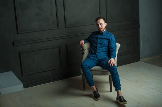 studio portrait of a young man on a dark wall background in a blue shirt and jeans. hipster man, dressed stylishly, with a beard and sunglasses, sitting in a light easy chair as a boss