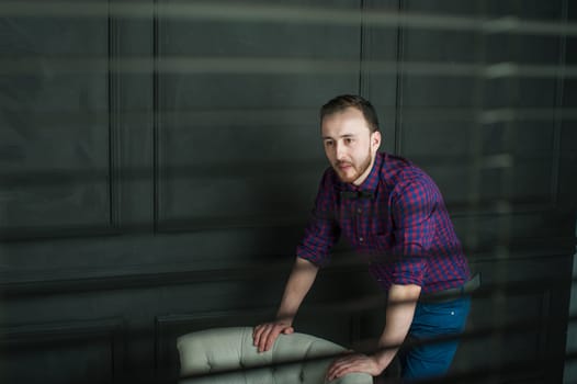 studio portrait of a young man on a dark wall background in a burgundy plaid shirt and blue jeans. hipster man, dressed stylishly, with a beard, in a room with an armchair