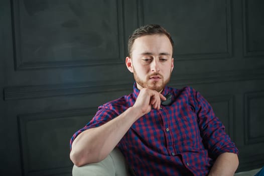 studio portrait of a young man on a dark wall background in a burgundy plaid shirt and blue jeans. hipster man, dressed stylishly, with a beard, in a room with an armchair