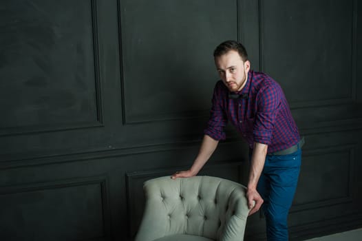 studio portrait of a young man on a dark wall background in a burgundy plaid shirt and blue jeans. hipster man, dressed stylishly, with a beard, in a room with an armchair