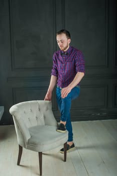 studio portrait of a young man on a dark wall background in a burgundy plaid shirt and blue jeans. hipster man, dressed stylishly, with a beard, in a room with an armchair