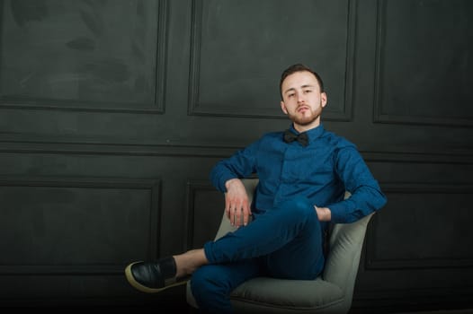 studio portrait of a young man on a dark wall background in a blue shirt and jeans. hipster man, dressed stylishly, with a beard, sitting in a light easy chair
