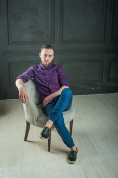 studio portrait of a young man on a dark wall background in a burgundy plaid shirt and blue jeans. hipster man, dressed stylishly, with a beard, in a room with an armchair