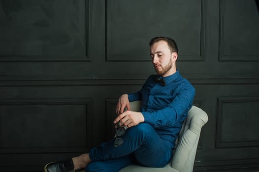 studio portrait of a young man on a dark wall background in a blue shirt and jeans. hipster man, dressed stylishly, with a beard and sunglasses, sitting in a light easy chair as a boss
