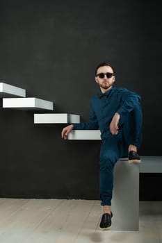 studio portrait of a young man on a dark wall background in a blue shirt and jeans. hipster man, dressed stylishly, with a beard and glasses, sitting on white stairs