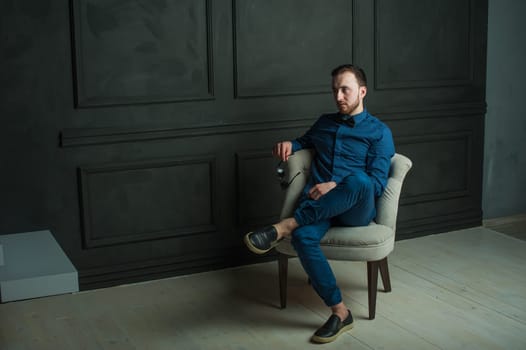 studio portrait of a young man on a dark wall background in a blue shirt and jeans. hipster man, dressed stylishly, with a beard and sunglasses, sitting in a light easy chair as a boss