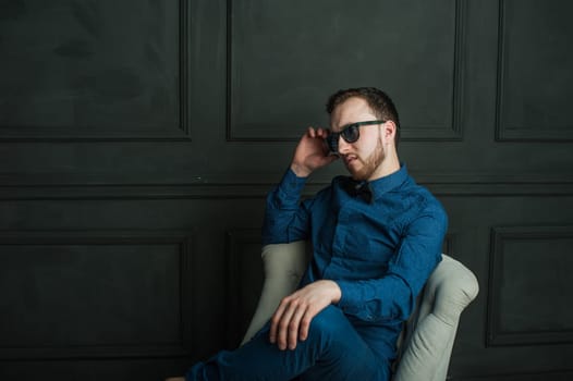 studio portrait of a young man on a dark wall background in a blue shirt and jeans. hipster man, dressed stylishly, with a beard and sunglasses, sitting in a light easy chair as a boss