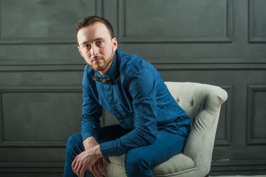 studio portrait of a young man on a dark wall background in a blue shirt and jeans. hipster man, dressed stylishly, with a beard, sitting in a light easy chair