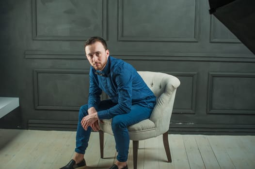 studio portrait of a young man on a dark wall background in a blue shirt and jeans. hipster man, dressed stylishly, with a beard, sitting in a light easy chair