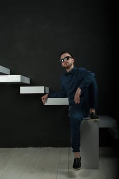studio portrait of a young man on a dark wall background in a blue shirt and jeans. hipster man, dressed stylishly, with a beard and glasses, sitting on white stairs