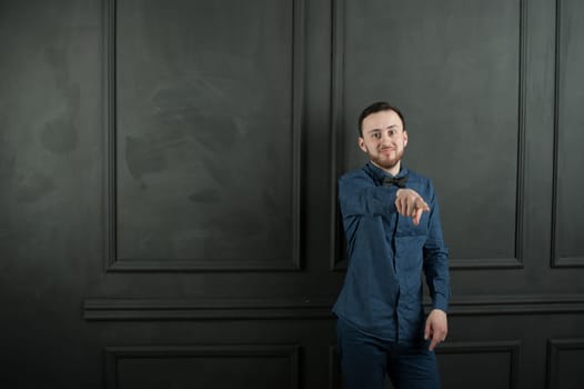 studio portrait of a young man on a dark wall background in a blue shirt and jeans. hipster man, dressed stylishly, with a beard, smiling and pointing finger