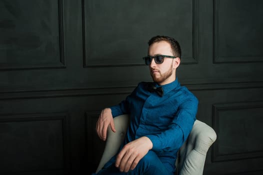 studio portrait of a young man on a dark wall background in a blue shirt and jeans. hipster man, dressed stylishly, with a beard and sunglasses, sitting in a light easy chair as a boss