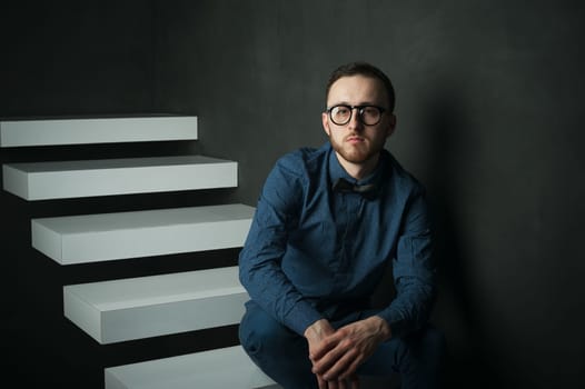 studio portrait of a young man on a dark wall background in a blue shirt and jeans. hipster man, dressed stylishly, with a beard and glasses, sitting on white stairs