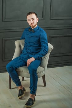 studio portrait of a young man on a dark wall background in a blue shirt and jeans. hipster man, dressed stylishly, with a beard, sitting in a light easy chair