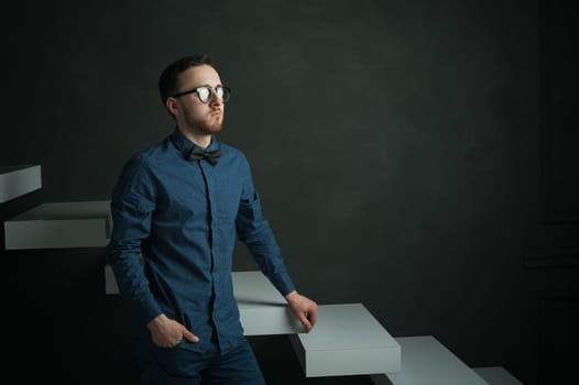 studio portrait of a young man on a dark wall background in a blue shirt and jeans. hipster man, dressed stylishly, with a beard and glasses, sitting on white stairs