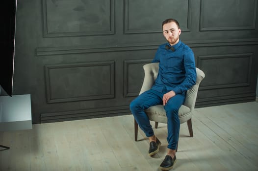 studio portrait of a young man on a dark wall background in a blue shirt and jeans. hipster man, dressed stylishly, with a beard, sitting in a light easy chair