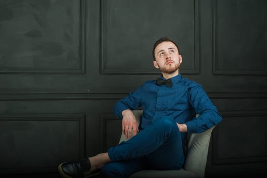 studio portrait of a young man on a dark wall background in a blue shirt and jeans. hipster man, dressed stylishly, with a beard, sitting in a light easy chair