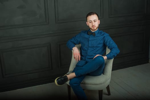 studio portrait of a young man on a dark wall background in a blue shirt and jeans. hipster man, dressed stylishly, with a beard, sitting in a light easy chair